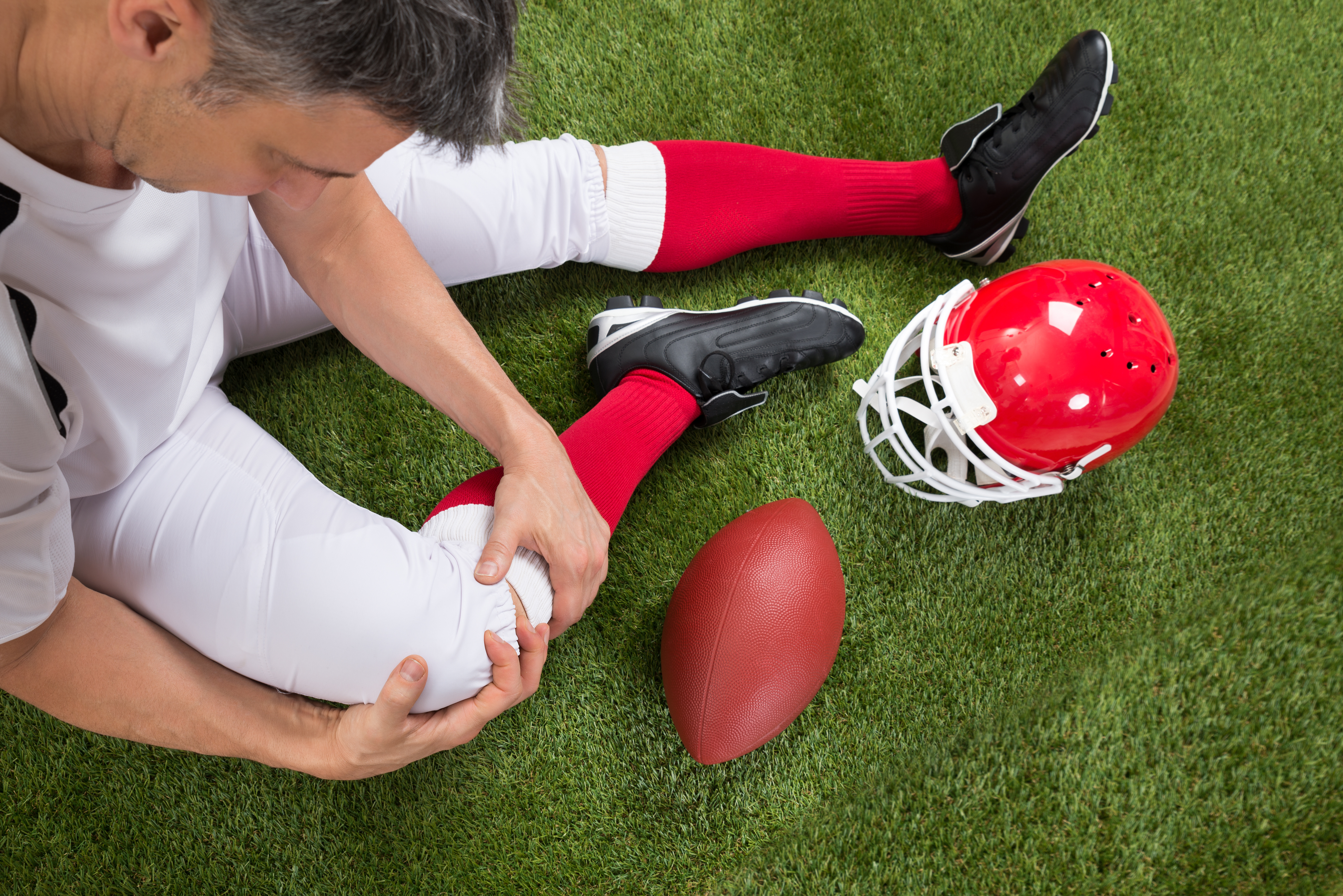 Football player injured on a green field, holding his knee. A helmet and football are nearby.
