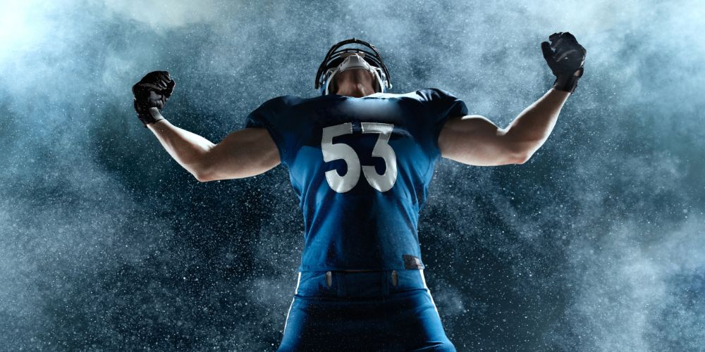 A football player wearing a blue jersey with the number 53 on it is standing in the rain with his arms outstretched.